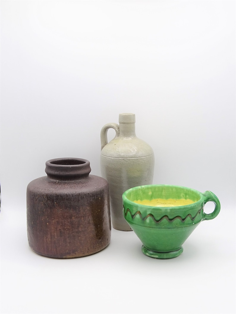 May include: Three ceramic vessels: a brown, cylindrical jar; a tall, off-white jug with a handle; and a green cup with a yellow interior and a decorative wavy brown line. The objects are arranged on a white background.