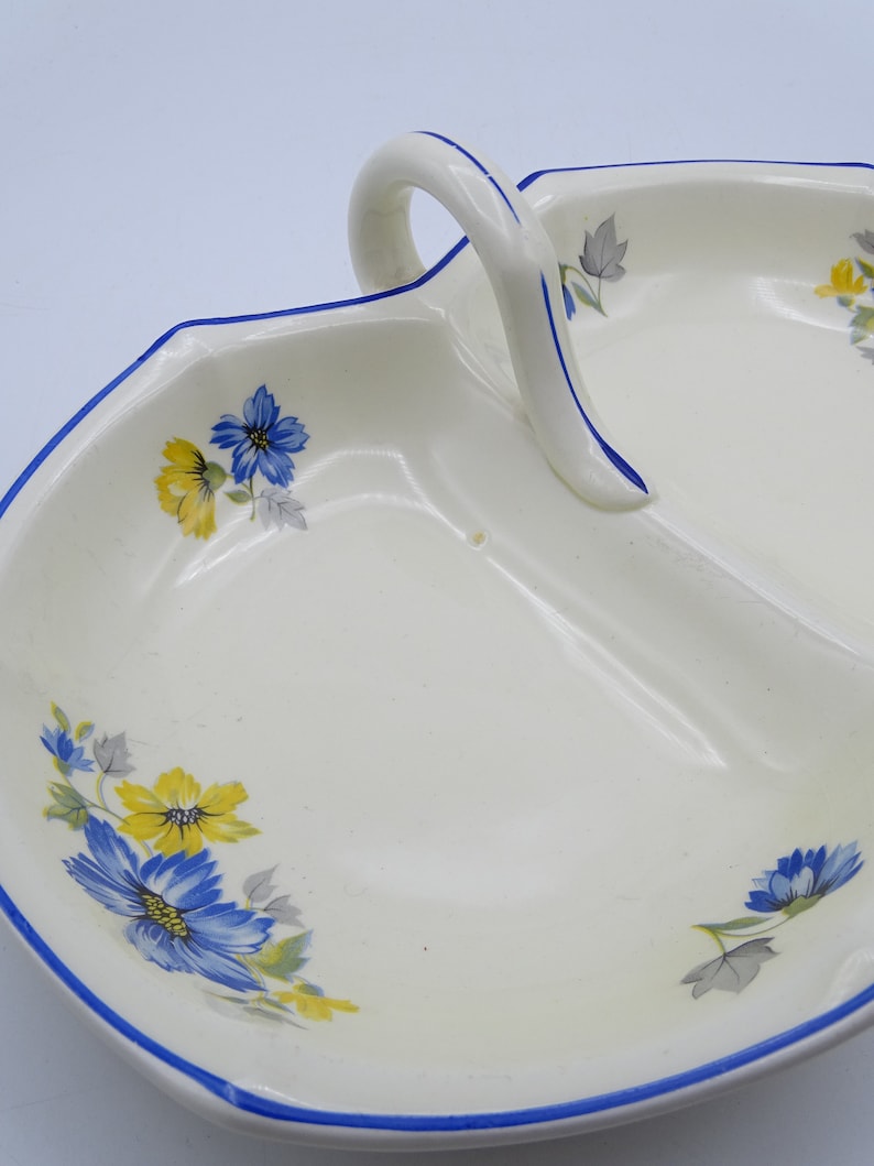 Może przedstawiać: Biała ceramiczna miska do serwowania z niebieskim i ż&oacute;łtym wzorem kwiatowym. Miska ma dwie sekcje i uchwyt.