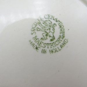 Może przedstawiać: Zielony i biały talerz ceramiczny z zieloną pieczątką, na kt&oacute;rej widnieje napis "Made in Holland" oraz lew z koroną i napis "Ceramique Maastricht".