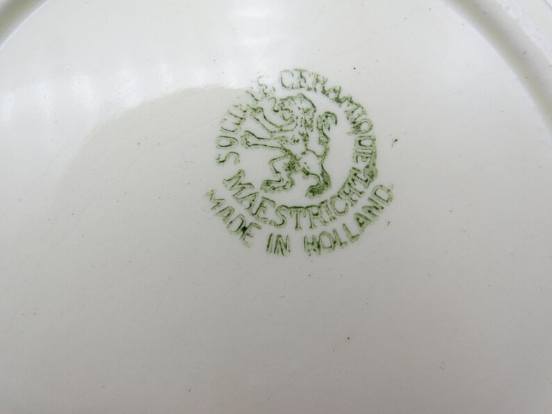 Może przedstawiać: Zielony i biały talerz ceramiczny z zieloną pieczątką, na kt&oacute;rej widnieje napis "Made in Holland" oraz lew z koroną i napis "Ceramique Maastricht".