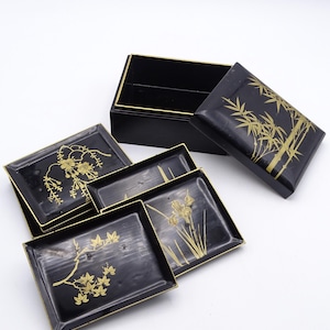 May include: A black lacquered box with a gold trim and floral designs. The box is open and contains four smaller black lacquered trays with gold trim and floral designs.