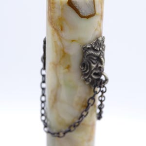 May include: A decorative bottle stopper with a cylindrical body made of cream and brown marbled stone. The stopper has a silver-colored metal top and two ornate metal faces with chains attached to the body.