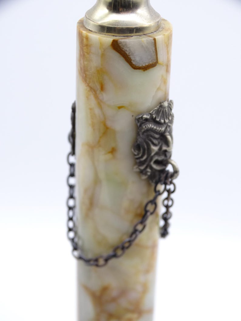 May include: A decorative bottle stopper with a cylindrical body made of cream and brown marbled stone. The stopper has a silver-colored metal top and two ornate metal faces with chains attached to the body.