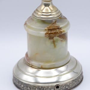 May include: A decorative lamp with a silver base and accents. The lamp's body is made of a light green and brown onyx material. The base has a detailed floral pattern around the bottom edge. The lamp is a unique home decor item.