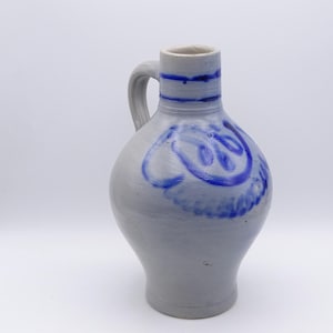 Puede incluir: Una jarra de gres azul y blanco con asa. La jarra tiene un diseño azul en la parte delantera.
