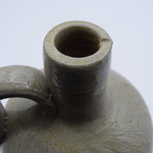 May include: Close-up of a stoneware jug with a handle. The jug is a light gray color with a textured surface and a rounded body. The opening is a dark brown color.