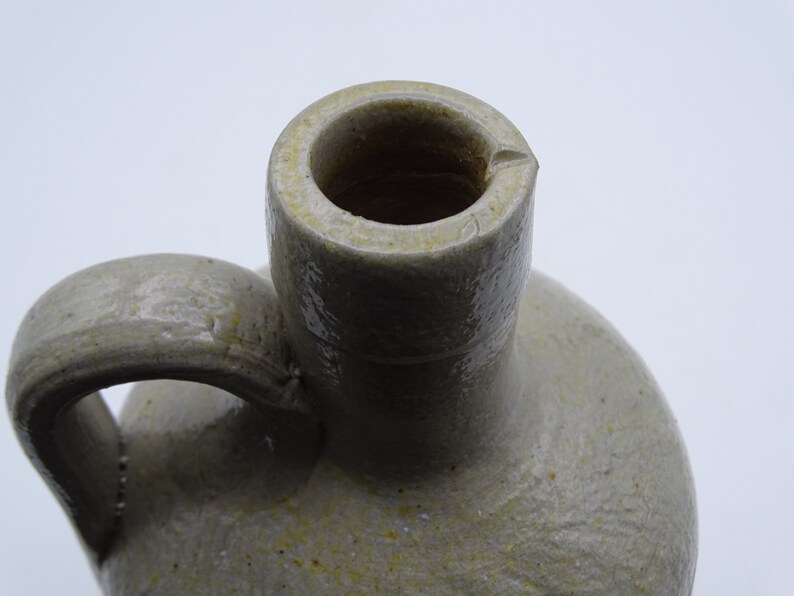 May include: Close-up of a stoneware jug with a handle. The jug is a light gray color with a textured surface and a rounded body. The opening is a dark brown color.