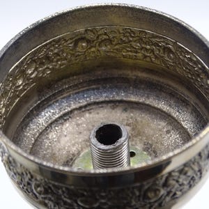 May include: Close-up of a silver-colored metal lampshade component. The lampshade features an ornate floral design around the rim and a threaded central post. The interior has a textured, aged appearance.
