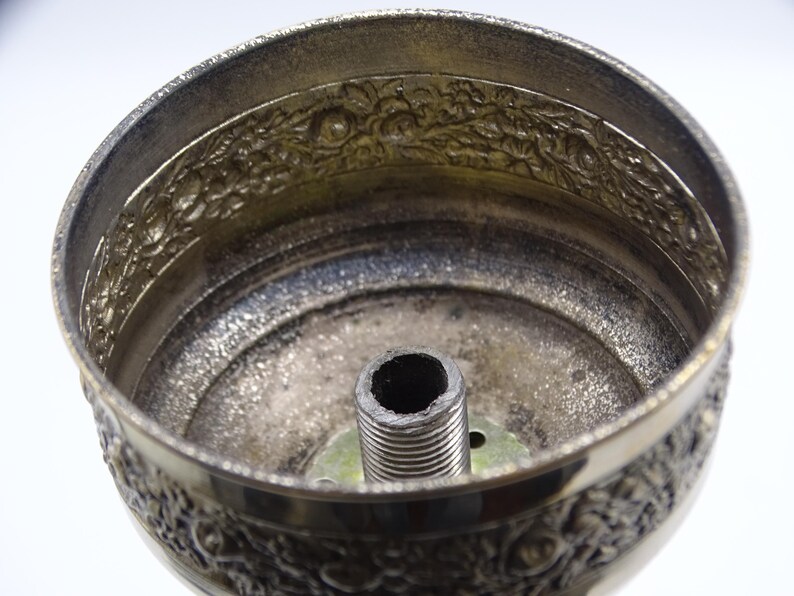 May include: Close-up of a silver-colored metal lampshade component. The lampshade features an ornate floral design around the rim and a threaded central post. The interior has a textured, aged appearance.