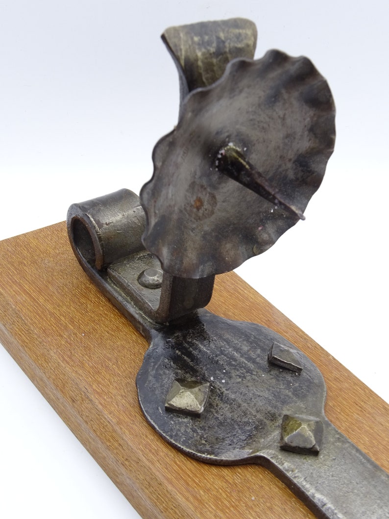 May include: A rustic, black metal candle holder with a scalloped top and a pointed spike for holding a candle. The holder is mounted on a wooden base.
