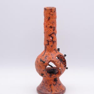Puede incluir: Un jarrón de cerámica naranja con una superficie texturizada y una forma distintiva. El jarrón presenta un cuello cilíndrico, una sección central bulbosa con una abertura circular y una base acampanada. Tiene detalles en negro y una textura rugosa y porosa. El diseño es artístico.