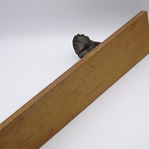 May include: A wooden shelf with a metal bracket in the shape of a seashell. The shelf is light brown and the bracket is dark gray.