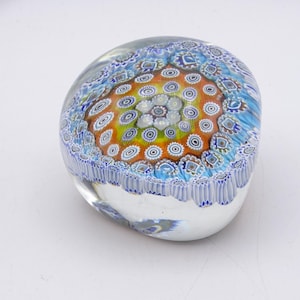 May include: A clear glass paperweight with a colorful millefiori design. The center features a pattern of orange, yellow, and white circles, surrounded by blue and white floral patterns. The base is clear, and the overall shape is rounded.