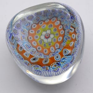 May include: A clear glass paperweight with a colorful, intricate design. The design features a central orange and yellow pattern surrounded by blue and white floral motifs. The paperweight is approximately 4 inches wide.