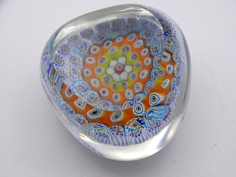 May include: A clear glass paperweight with a colorful, intricate design. The design features a central orange and yellow pattern surrounded by blue and white floral motifs. The paperweight is approximately 4 inches wide.