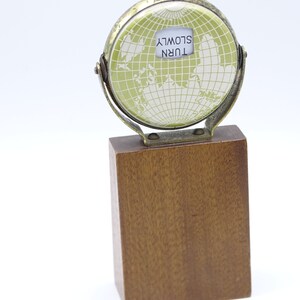 May include: A small, brass globe with a green map and a white label that reads "TURN SLOWLY". The globe is mounted on a wooden base.