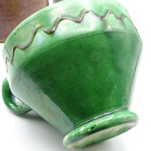 May include: A green ceramic bowl with a wavy, gold-colored trim around the rim. The bowl has a handle and a footed base. The glaze has a glossy finish. The bowl is tilted, showing its interior and the underside of the base.