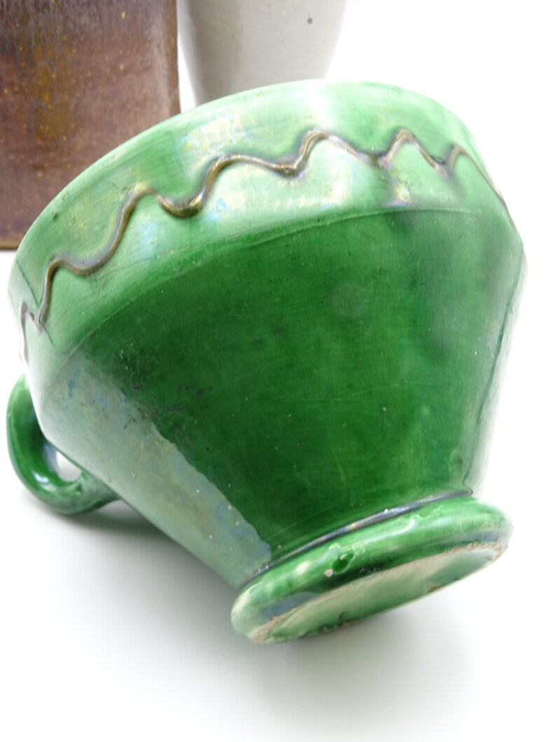 May include: A green ceramic bowl with a wavy, gold-colored trim around the rim. The bowl has a handle and a footed base. The glaze has a glossy finish. The bowl is tilted, showing its interior and the underside of the base.