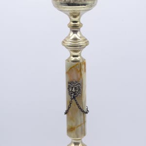 May include: A tall, ornate candlestick with a gold-colored metal top and base. The central column is made of a light green and brown stone, with a decorative lion's head and chain. The top has a floral design.