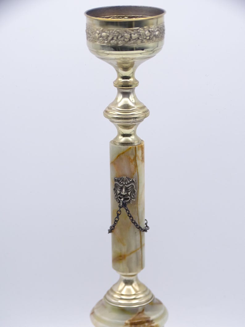 May include: A tall, ornate candlestick with a gold-colored metal top and base. The central column is made of a light green and brown stone, with a decorative lion's head and chain. The top has a floral design.