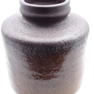 May include: A dark brown, cylindrical ceramic jar with a textured surface. The jar has a rounded body and a narrow, slightly flared neck. The color transitions from a lighter brown at the base to a darker, almost black, shade at the top. The jar is likely for storage or decoration.