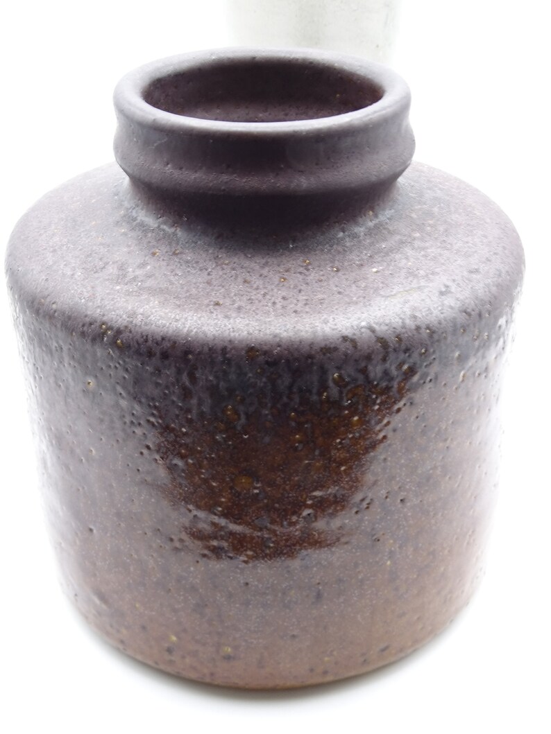 May include: A dark brown, cylindrical ceramic jar with a textured surface. The jar has a rounded body and a narrow, slightly flared neck. The color transitions from a lighter brown at the base to a darker, almost black, shade at the top. The jar is likely for storage or decoration.