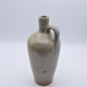 May include: A stoneware jug with a handle. The jug is a light brown color with a rounded body and a narrow neck. It has a handle on the side and a small opening at the top. The jug appears to be antique or vintage.