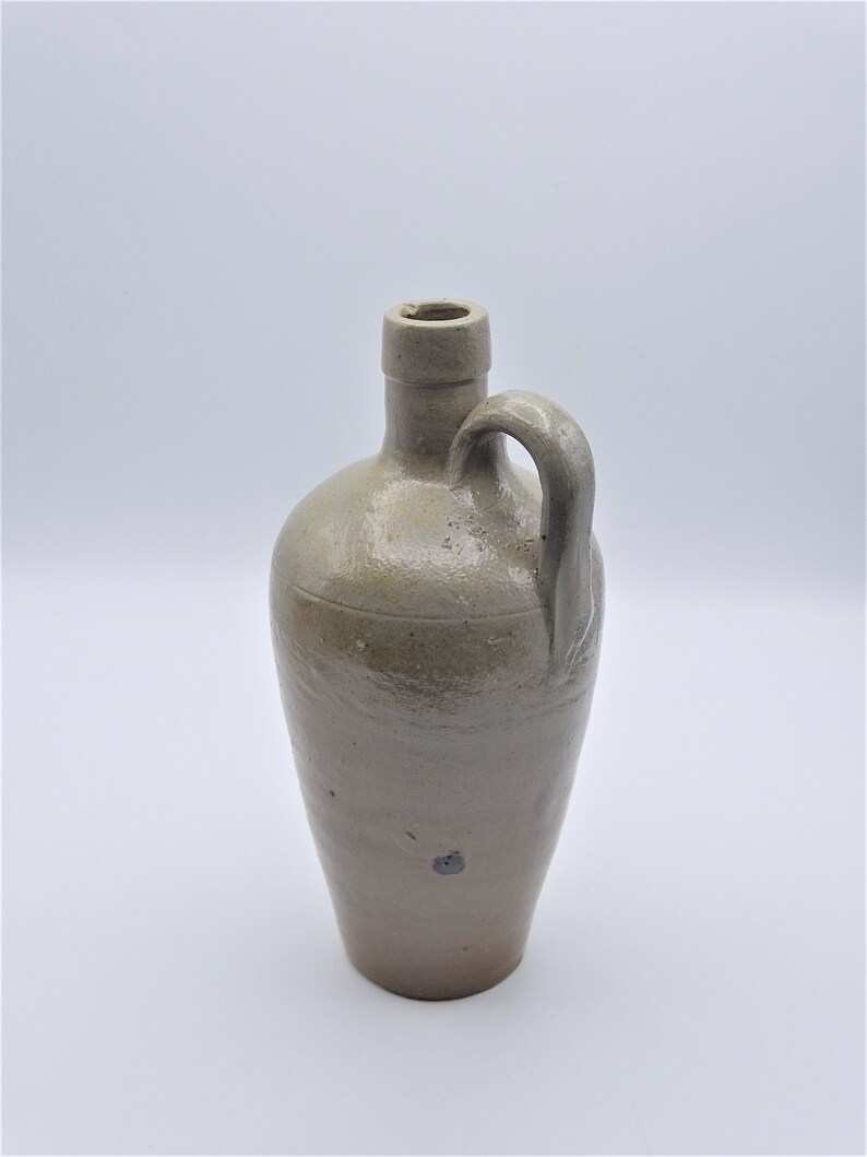 May include: A stoneware jug with a handle. The jug is a light brown color with a rounded body and a narrow neck. It has a handle on the side and a small opening at the top. The jug appears to be antique or vintage.