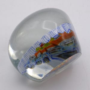 May include: A clear glass paperweight with a rounded shape. The interior features a colorful design with orange, blue, and green accents. A white and blue striped pattern borders the design, adding a decorative touch.