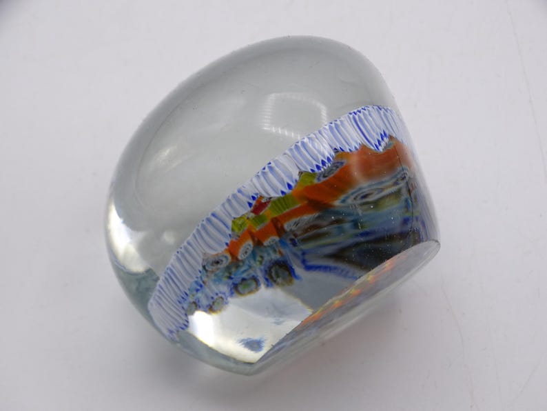 May include: A clear glass paperweight with a rounded shape. The interior features a colorful design with orange, blue, and green accents. A white and blue striped pattern borders the design, adding a decorative touch.