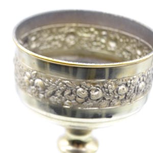 May include: A silver-colored goblet with a detailed floral pattern around the bowl. The rim has a gold accent. The goblet sits on a pedestal base. The design is ornate and vintage.