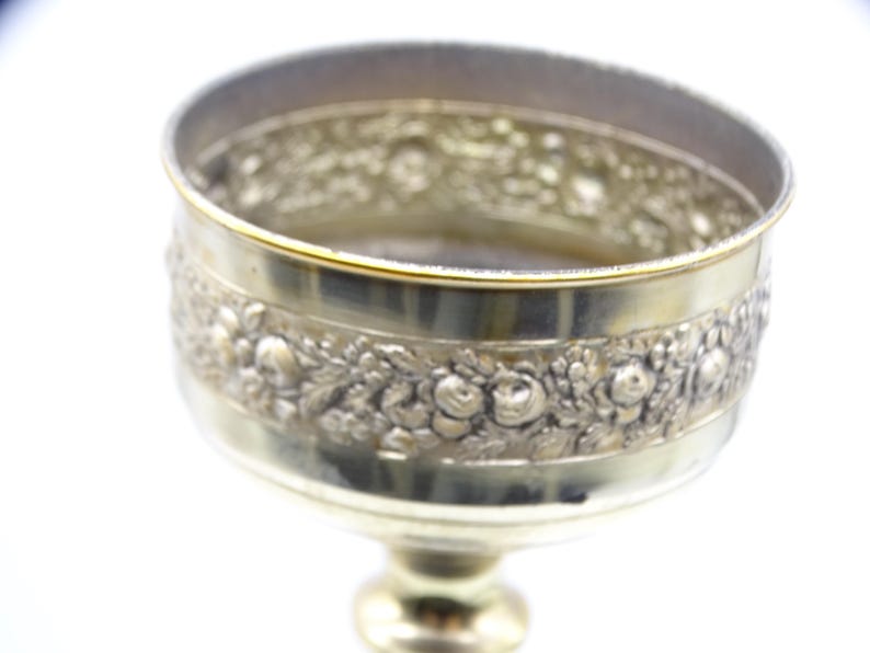 May include: A silver-colored goblet with a detailed floral pattern around the bowl. The rim has a gold accent. The goblet sits on a pedestal base. The design is ornate and vintage.