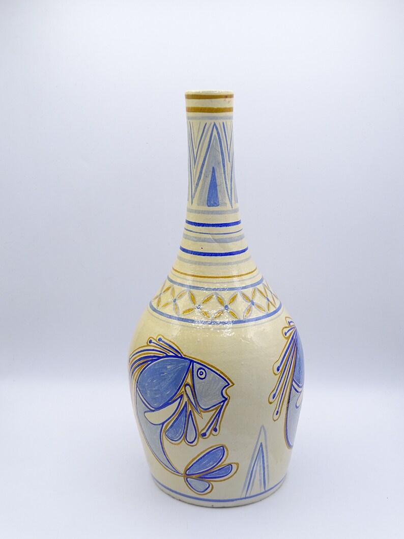 May include: A ceramic vase with a light yellow background and blue and gold painted fish designs. The vase has a narrow neck and a wide, rounded body.