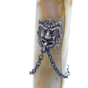 May include: Close-up of a decorative pen with a silver-colored face charm and chain. The pen's body has a cream and brown marbled pattern. The charm is a detailed, stylized face with a chain attached.