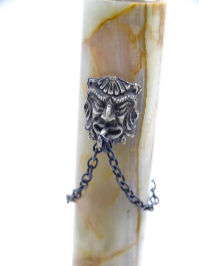 May include: Close-up of a decorative pen with a silver-colored face charm and chain. The pen's body has a cream and brown marbled pattern. The charm is a detailed, stylized face with a chain attached.