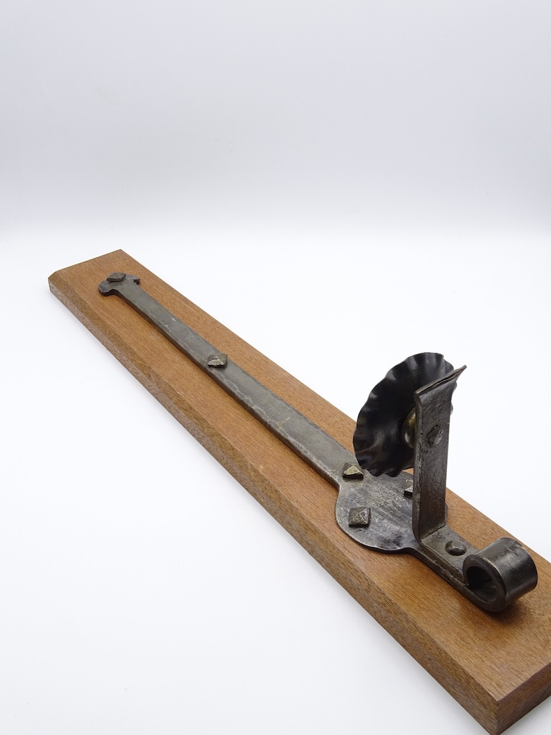 May include: A long, thin, metal hinge with a decorative wheel at one end. The hinge is mounted on a wooden board.
