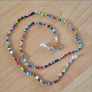 Può includere: Una catena per occhiali da vista in perline colorate con due chiusure argentate. La catena è realizzata con piccole perline rotonde di vari colori, tra cui rosso, blu, verde, giallo e nero.