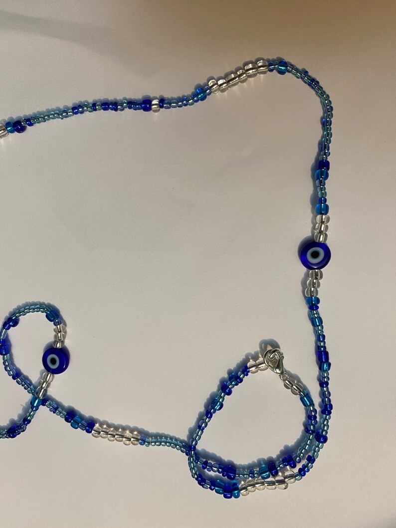 以下が含まれることがあります： 青いビーズと銀色のビーズでできたネックレスとブレスレットのセットで、邪眼のチャームが付いています。ネックレスはブレスレットよりも長く、どちらも銀色の留め具が付いています。