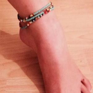 Op de afbeelding: Een close-up van een enkel met een meerkleurig kralen armbandje. Het armbandje is gemaakt van kleine, kleurrijke kralen en is drie keer om de enkel gewikkeld.