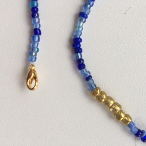 Pode incluir: Um colar de contas azuis e douradas com um fecho dourado. O colar &eacute; feito de pequenas contas redondas em tons de azul, com uma se&ccedil;&atilde;o de contas douradas no centro.