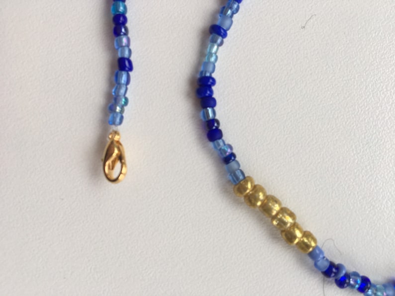 Pode incluir: Um colar de contas azuis e douradas com um fecho dourado. O colar &eacute; feito de pequenas contas redondas em tons de azul, com uma se&ccedil;&atilde;o de contas douradas no centro.