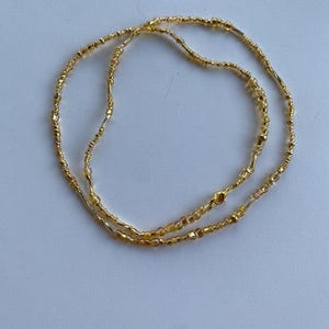 Könnte beinhalten: Eine goldene Perlen-Taillenkette mit einem Verschluss. Die Kette besteht aus kleinen, runden Perlen und einigen größeren, rechteckigen Perlen.
