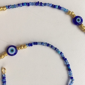 Pode incluir: Um colar de contas azuis e douradas com dois pingentes de olho turco azul. O colar &eacute; feito com contas pequenas e redondas em tons de azul e dourado.