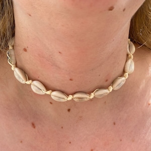 May include: A simple, minimalist choker necklace made with natural seashells and a cream-coloured cord. The necklace is tied with a knot at the back of the neck.
