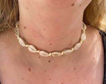 May include: A simple, minimalist choker necklace made with natural seashells and a cream-coloured cord. The necklace is tied with a knot at the back of the neck.