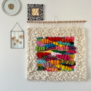 May include: A colorful woven wall hanging with a white background and a variety of bright colors, including red, orange, yellow, green, blue, purple, and pink. The hanging is made with different textures and patterns, creating a unique and eye-catching piece of art.