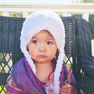 May include: A young child wearing a white knitted hat with braids, a purple shawl, and a white embroidered blouse with colorful accents.