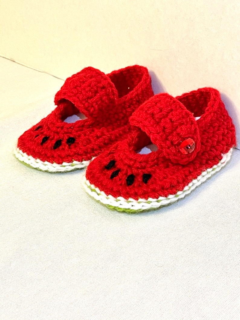Puede incluir: Un par de zapatos de beb&eacute; de crochet rojo con un dise&ntilde;o de sand&iacute;a. Los zapatos tienen un borde blanco y un borde verde alrededor de la parte inferior.
