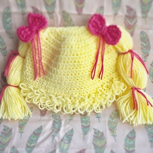 Puede incluir: Un gorro de crochet amarillo con dos coletas de hilo amarillo y dos lazos de crochet rosa.