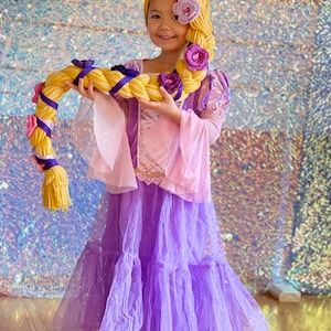 Könnte beinhalten: Ein junges Mädchen verkleidet als Rapunzel mit einem langen blonden Zopf, das ein lila Kleid mit einem rosa Oberteil und einer Blumenkrone trägt.